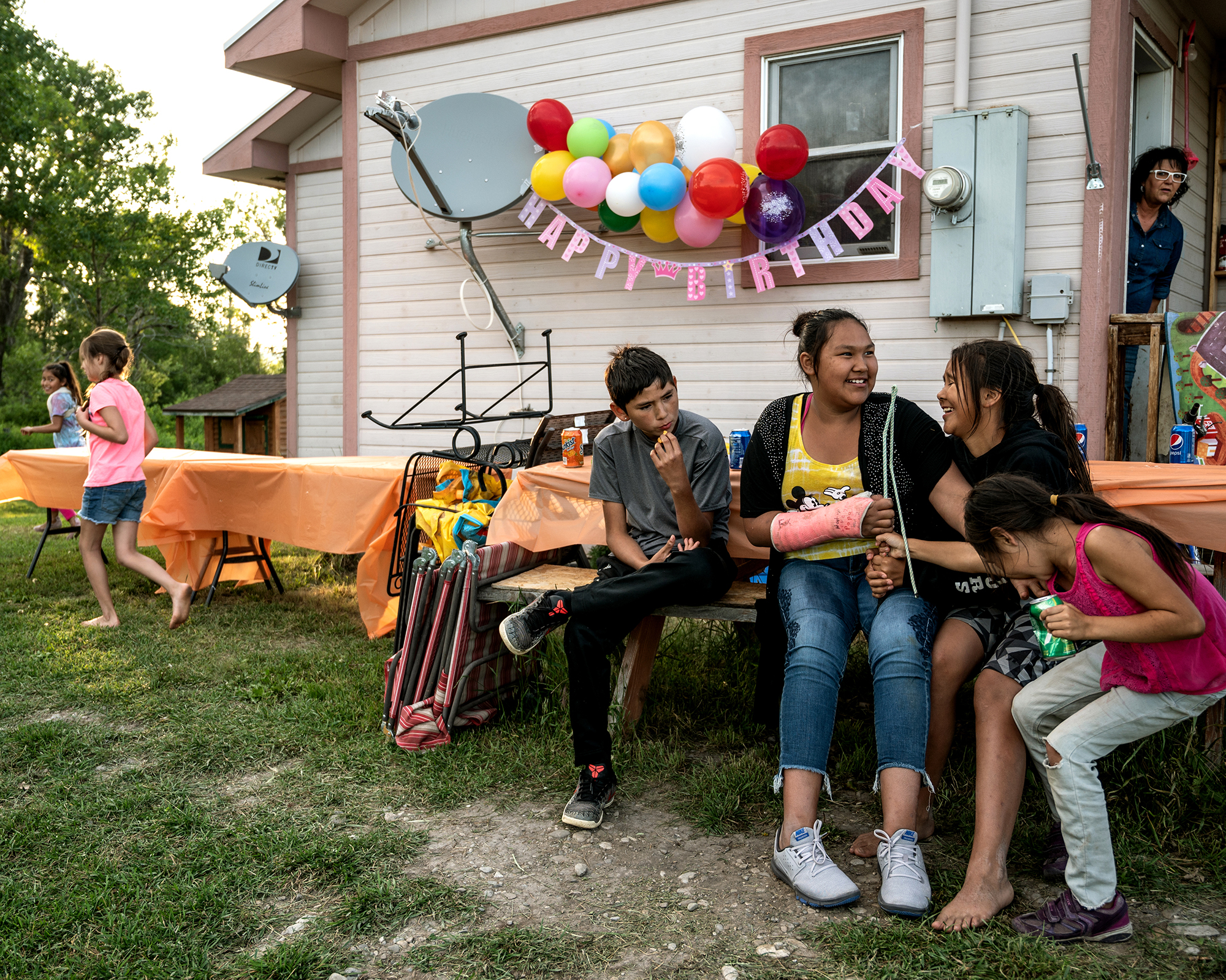 Four kids sit on a picnic table bench, laughing, with a "Happy Birthday" banner and balloons hanging off the house in the background.