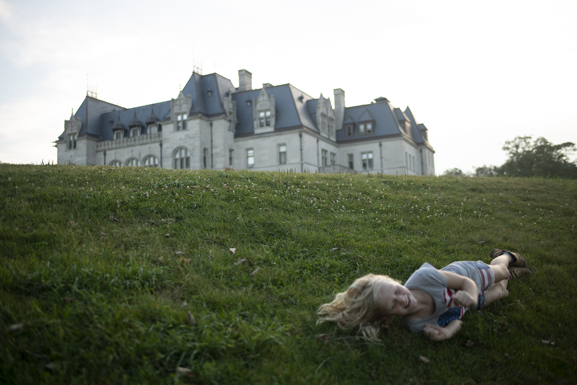 Young girl rolls down a grassy hill with a large mansion in the background.