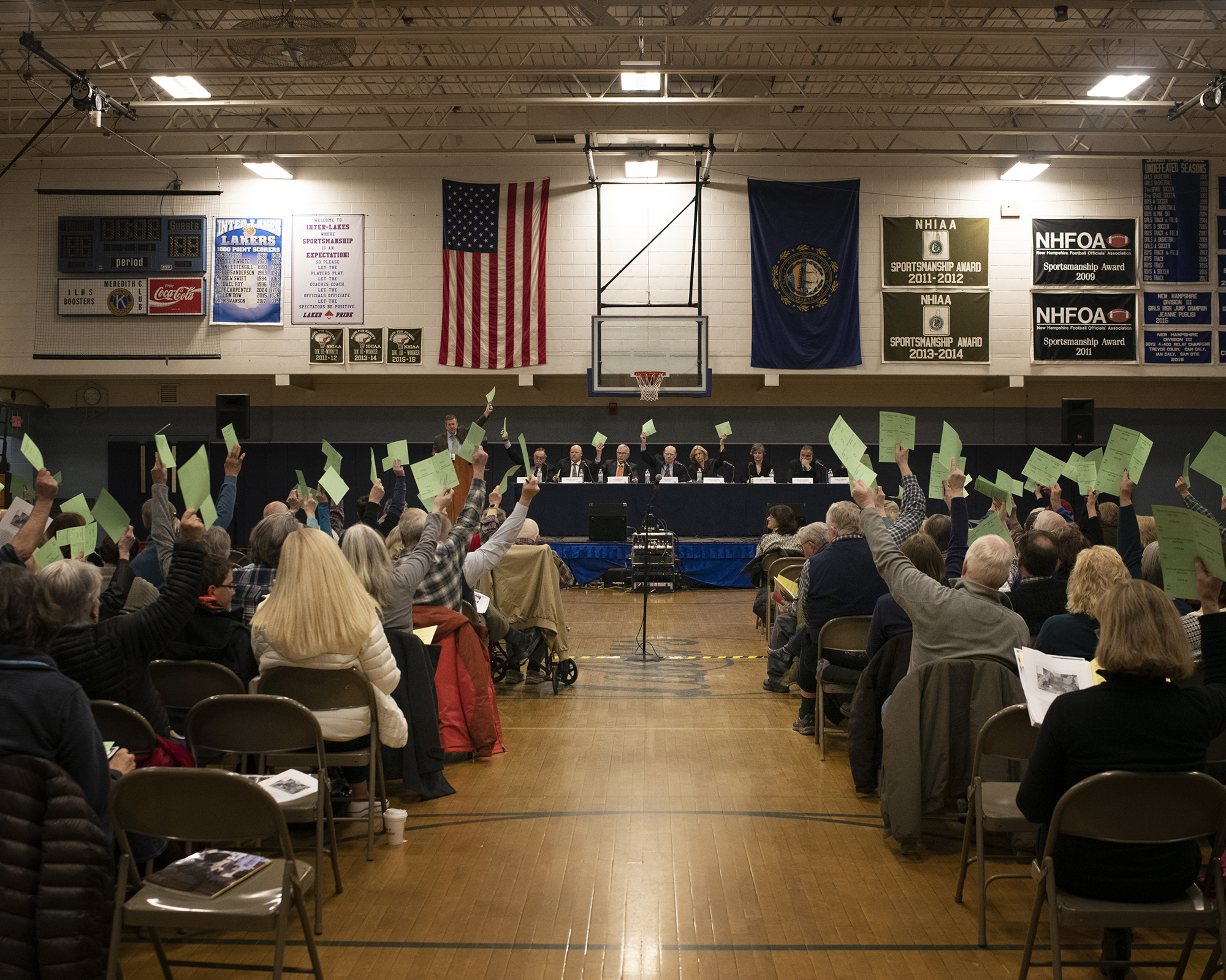 School gymnasium filled with people sitting in folding chairs, some with their arms raised, holding pieces of green paper.