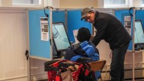 A white man explaining something to a person sitting in a chair at an accessible voting machine, with a walker next to the person in a chair.