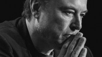 A closely cropped image of Elon Musk's face, as he leans forward, as his hands, pressed to his lips, steeple.