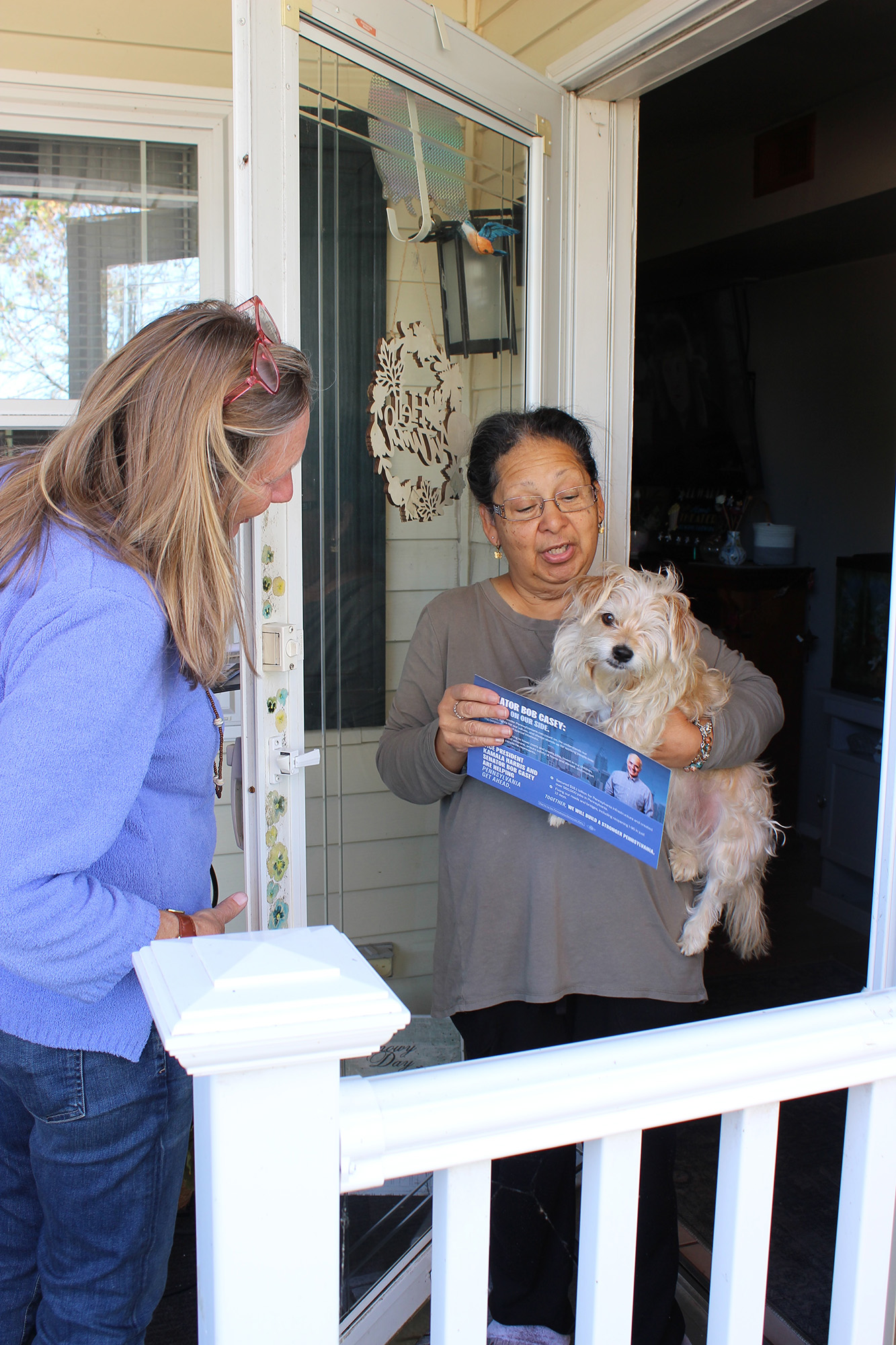 A blond woman talks to a woman in a doorway, holding a dog.