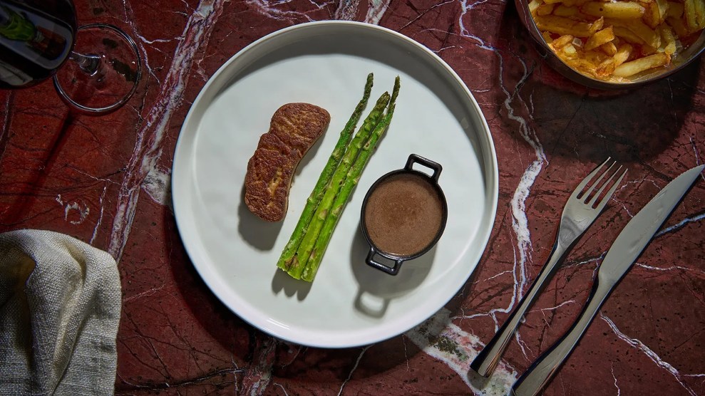 A white plate with faux foie gras, three asparagus, and a small caldron of brown liquid.