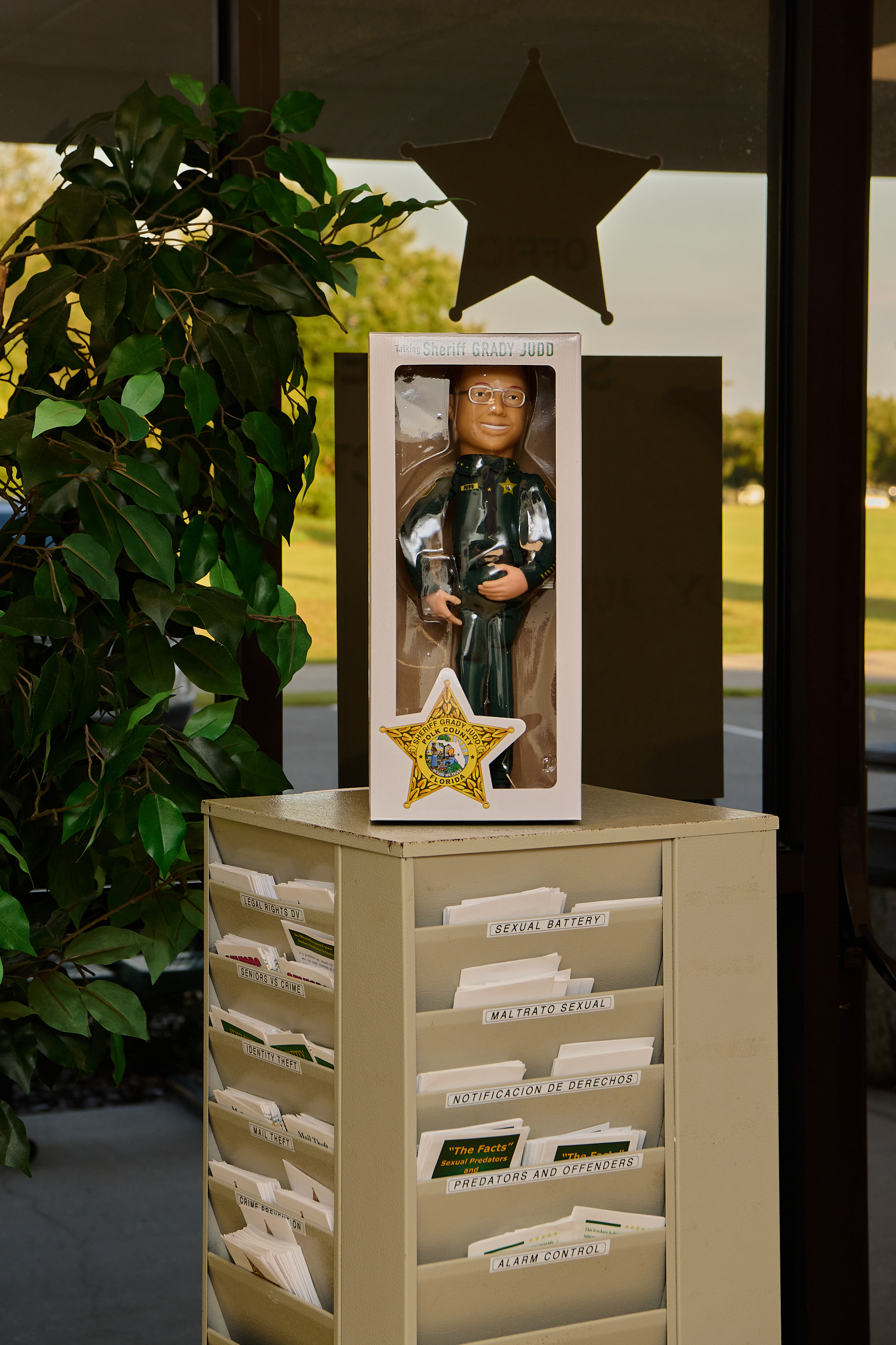 A toy doll of Sheriff Grady Judd in a plastic marketing box sits on top of a rack of pamphlets with the words "sexual battery."