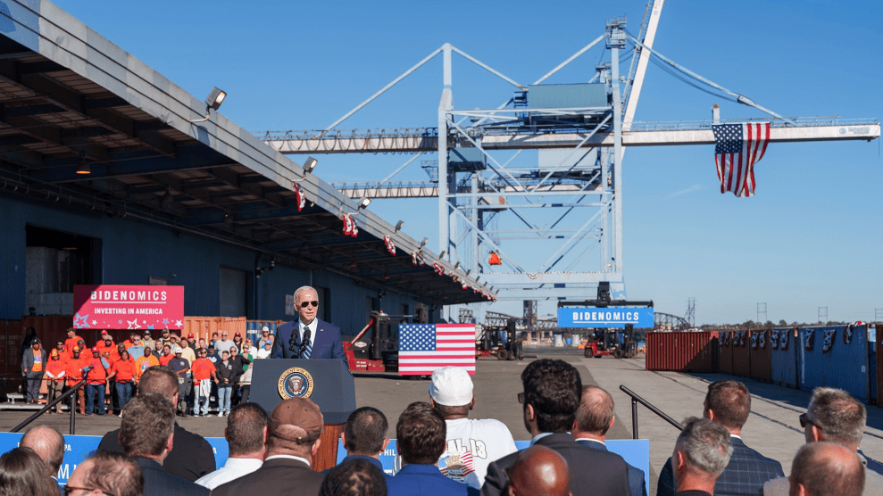 President Joe Biden speaks to a small crowd in front of a bridge. Several American flags flow behind him, in addition to signs that say "Bidenomics."