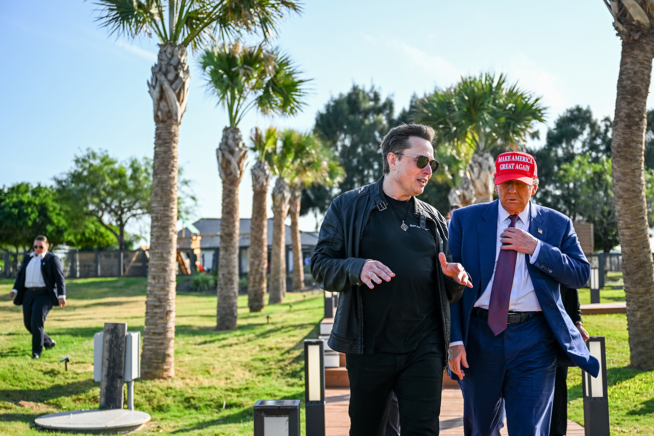 Elon Musk and Donald Trump, wearing a red "Make America Great Again" cap, talk while walking side by side.