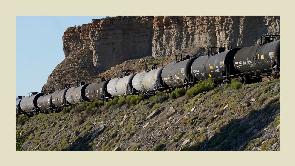 A line of tank cars in differing shades of black and gray stretch from one end of the photograph to the other. The tanks are part of a train that's traveling through rocky, brown mountainous terrain dotted with small green shrubs.