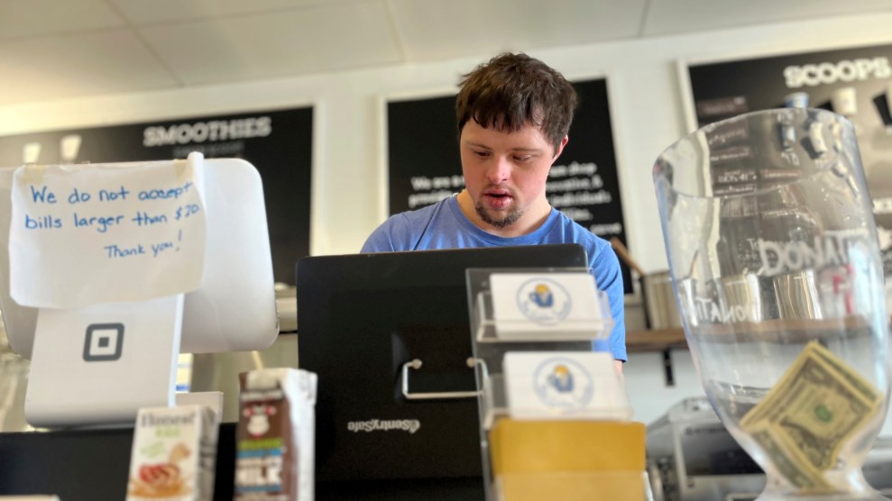 A white man with brown hair in a blue t-short in front of a black machine, taking an order. There are signs behind him that say "scoops" and smoothies."