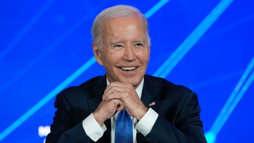 An older man (President Joe Biden) is smiling with one hand wrapped around the other that is in a fist. He is wearing a dark navy suit, white dress shirt, blue tie, and American flag pin. The background is a blue screen with white stripes.