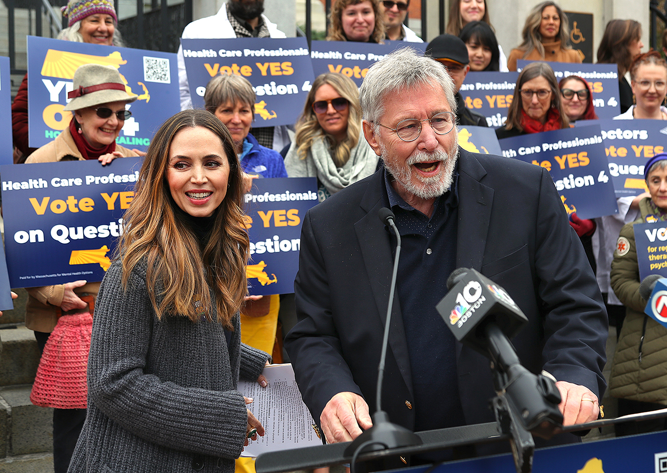 Eliza Dushku and Bessel van der Kolk stand near a microphone. Behind them, rows of people hold signs that read, "Health care professionals vote yes on Question 4."