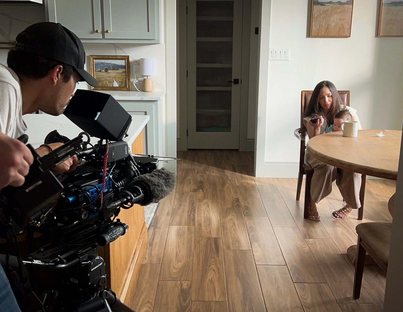 A man holding a large camera films a woman(Alejandra) while she speaks to her father on the phone and holds her child.