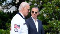 Hunter Biden wearing a suit looks at Joe Biden wearing a team USA jacker with a slight grin.