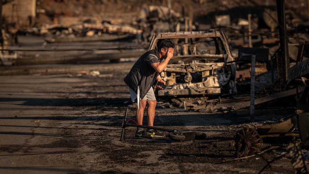 A young man in jacket and shorts stands next to his destroyed mobile home during the LA wildfires. Greiving, his right hand covers his face, as he holds his mobile phone in his left hand.