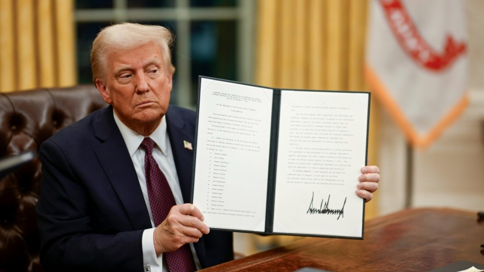 President Donald Trump sits at his desk in the Oval Office holding up a signed executive order.