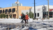 A group of three people bundled in jackets walk through an intersection covered in snow. The sky is blue behind them.