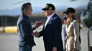 Two white men wearing suits shake hands. A woman in a tan jacket stands next to them.