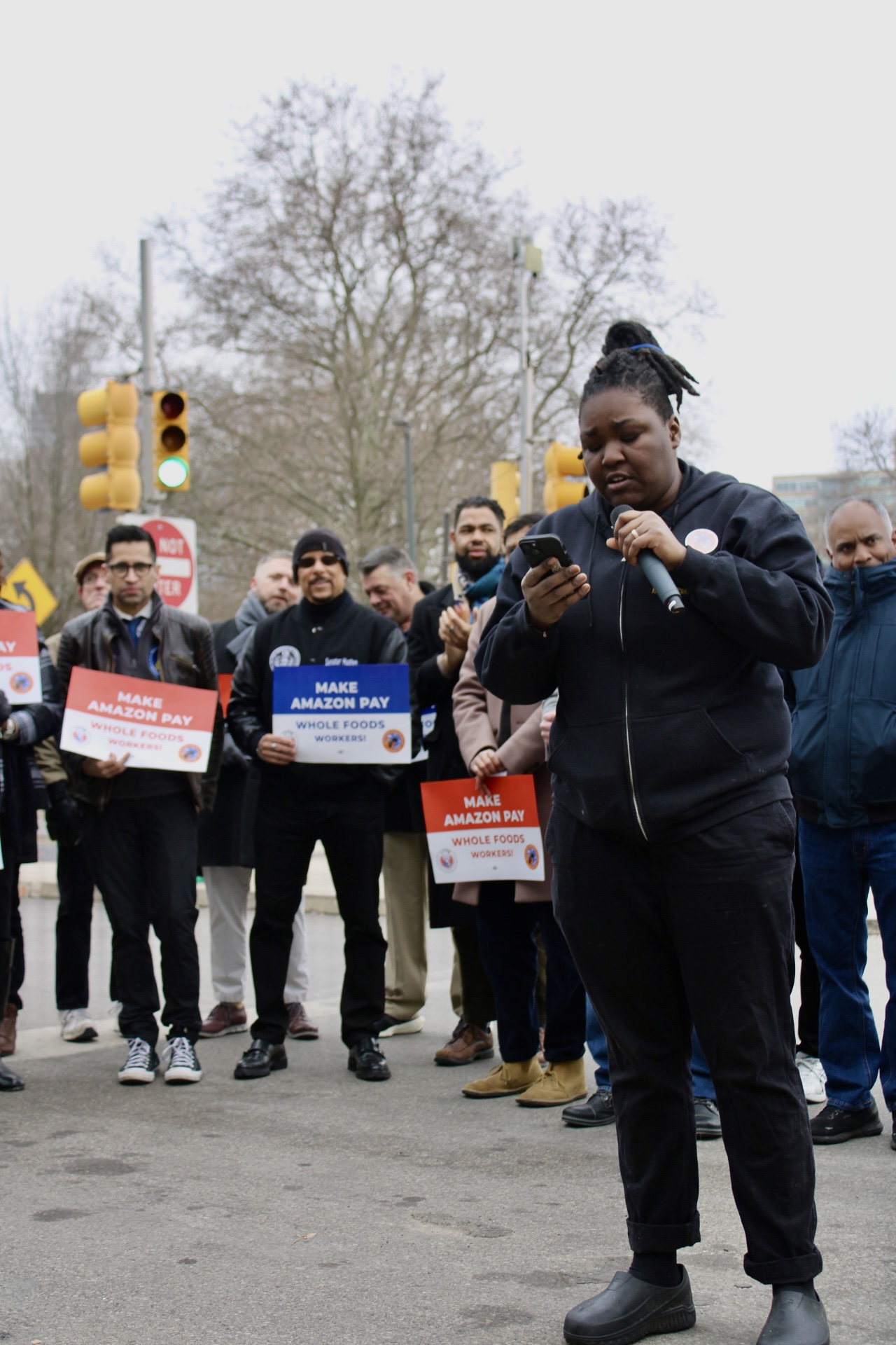 A woman speaks into a microphone. Behind her, an assembled group holds signs that read, "Make Amazon pay Whole Foods workers!" 