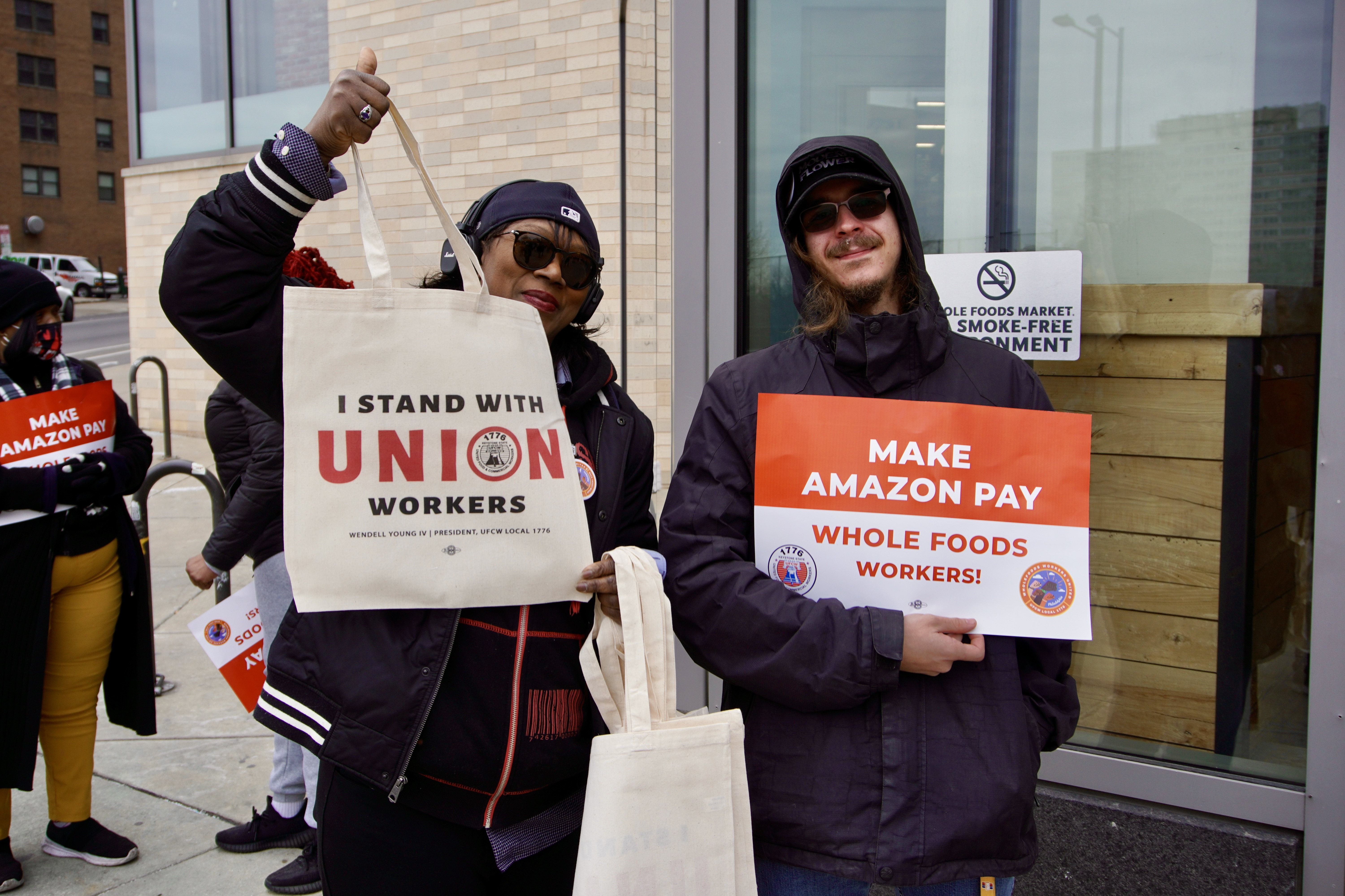 A woman holds up a tote bag that reads, "I stand with union workers." Next to her, a man holds a sign that reads, "Make Amazon pay Whole Foods workers!"