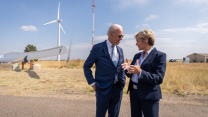 A white man and a white woman both wearing suits talk to each other in front of a windmill in a desert.