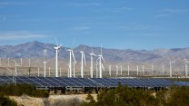 Solar panels and wind turbines in a desert in front of mountains.