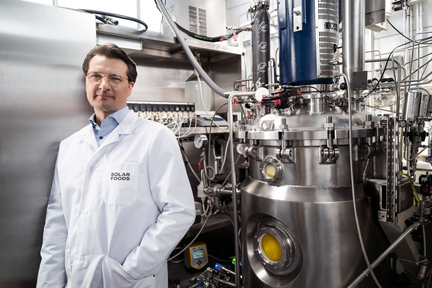 A white man in a white lab coat in front of metal machinery.