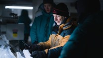 A man wearing a dirty yellow jacket looks at a polar ice core.