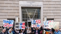 A crowd of people gather in front of an entrance to the USAID building. Some hold homemade signs that say things such as "Democracy Died in Complacency," "Save USAID, Save Lives," and "Musk & Trump: Keep Your Fascist Hands Off USAID."