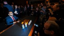 Kash Patel, dressed in dark blue suit, white shirt and red tie, sits a table behind a microphone and his name placard. Patel, who has a beard and wears thick -rimmed black glasses, is lit starkly by the flash of a camera. In front of him, a throng of photographers lean in for a close, detailed portrait.