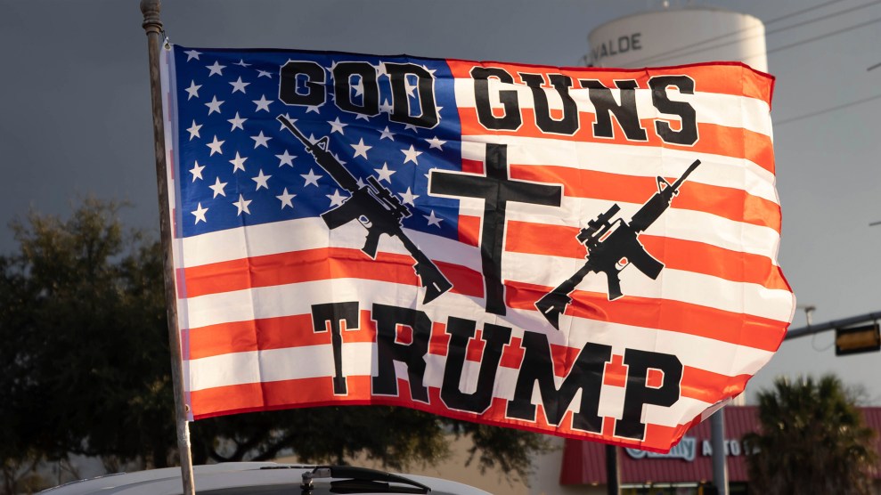 Large flag waves from a car, bearing the slogan "GOD, GUNS, TRUMP" with graphics of two rifles and a cross overlaid on the American flag stars and stripes.