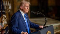 Donald Trump, wearing a navy suit and blue tie, stands behind a microphone.