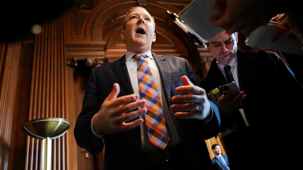 A white man wearing a plaid tie speaks with his mouth very wide open.