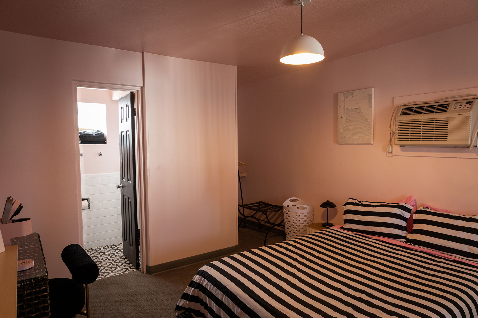 A basic hotel room with a wall-mounted air conditioner, a bed with striped bedding, and a small bathroom.