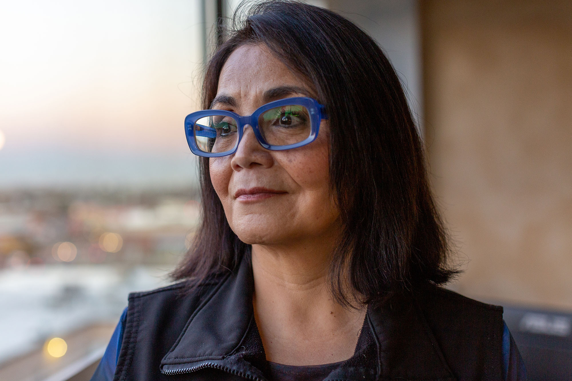 Portrait of woman by a window with thick-framed blue glasses.