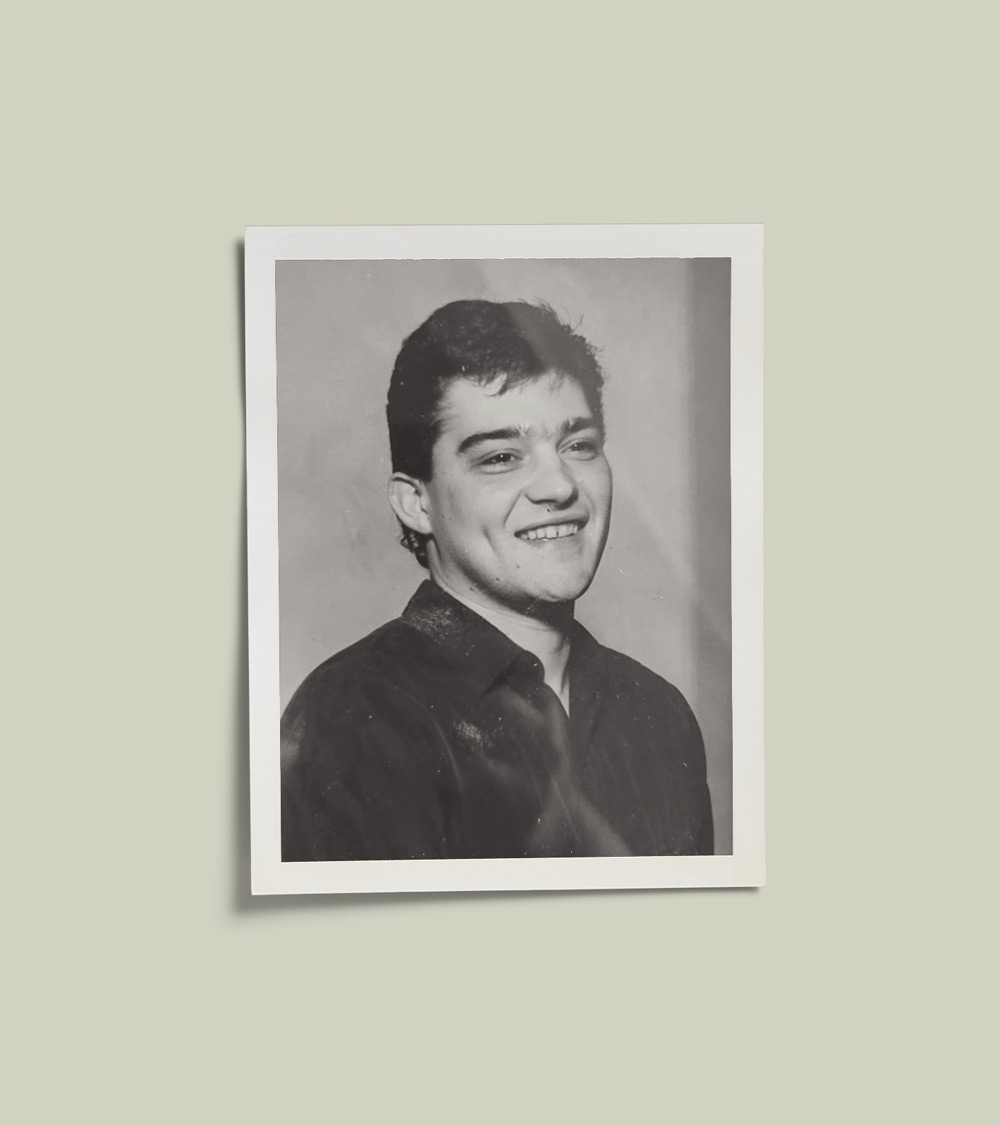 A black-and-white vintage photograph of a young man with short dark hair, wearing a collared shirt. He is smiling. The photo is set against a pale green background.