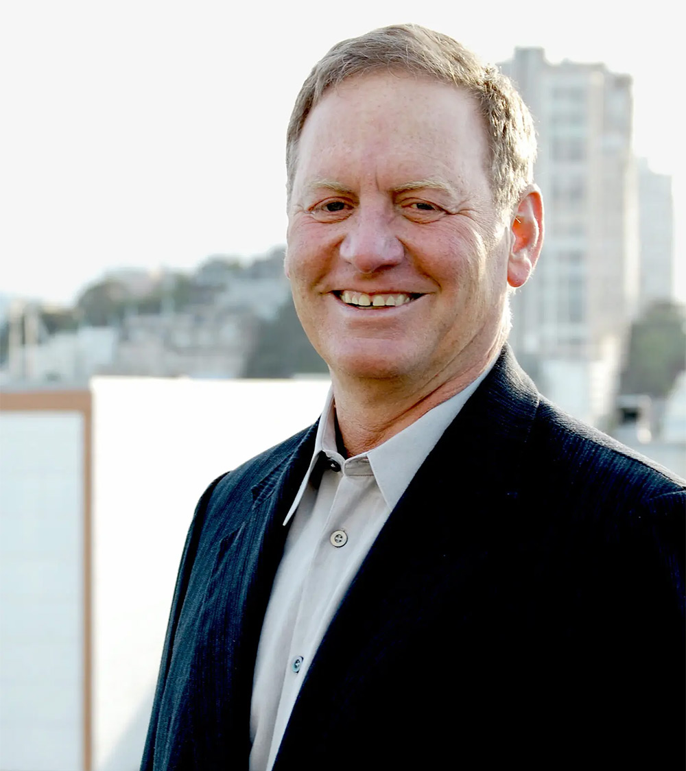 A middle-aged man smiling warmly outdoors, wearing a dark blazer and light-colored collared shirt. The background is softly blurred, showing urban buildings and greenery in the distance on a bright, clear day.