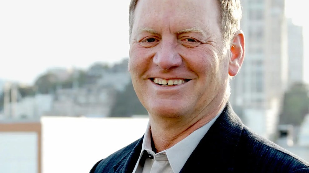 A middle-aged man smiling warmly outdoors, wearing a dark blazer and light-colored collared shirt. The background is softly blurred, showing urban buildings and greenery in the distance on a bright, clear day.