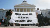 A photograph of a crowd of people assembled at the steps of the California state capitol building. In the foreground at the back of the crowd, are two young people holding a white sheet with words in black, all sans serif caps, that read: "HUMAN RIGHTS FOR PELICAN BAY PRISONERS NOW!"