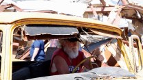 A man with a white beard drives a yellow truck in the middle of a destroyed landscape.