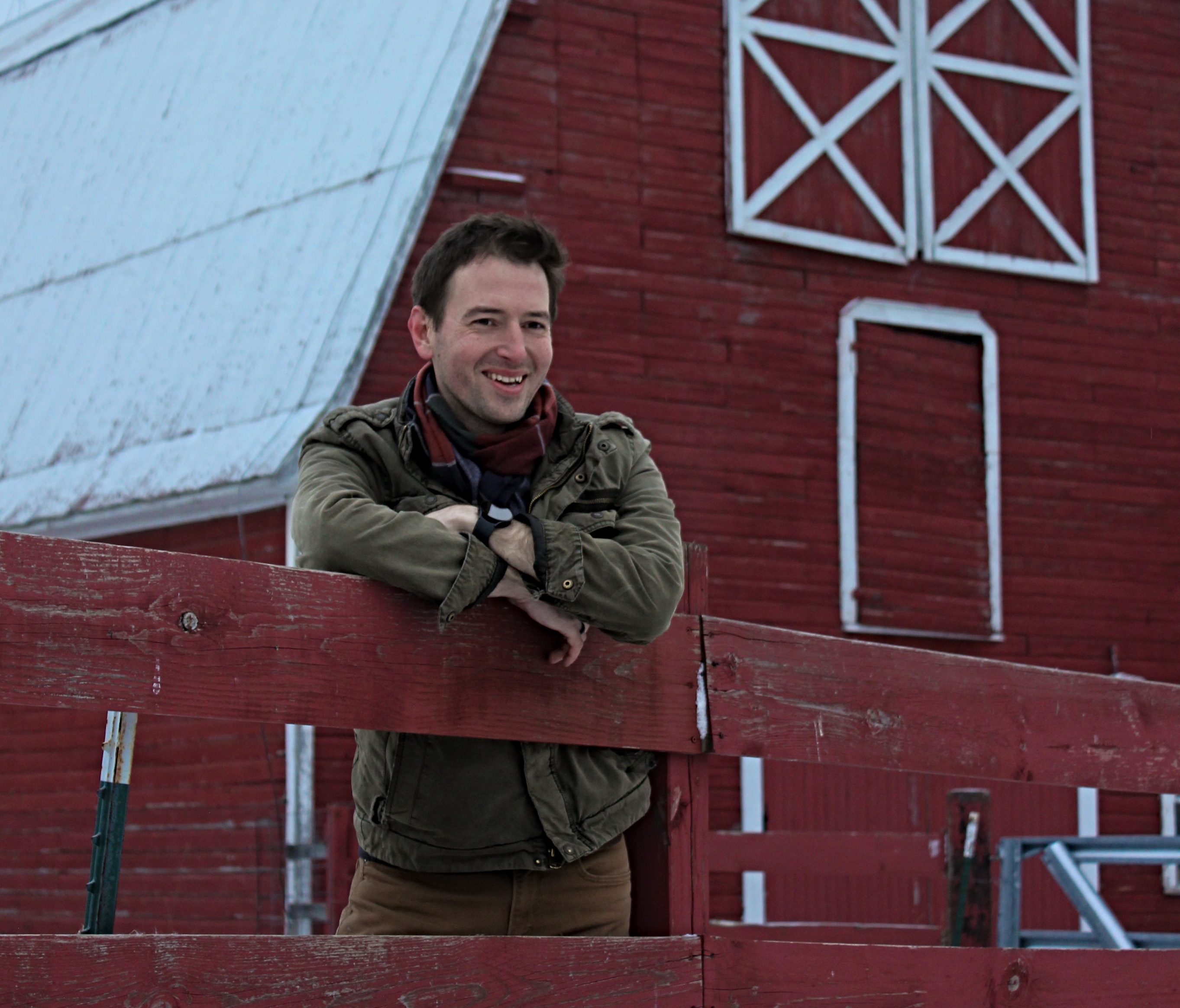 A man smiles while standing in front of a snowy red barn