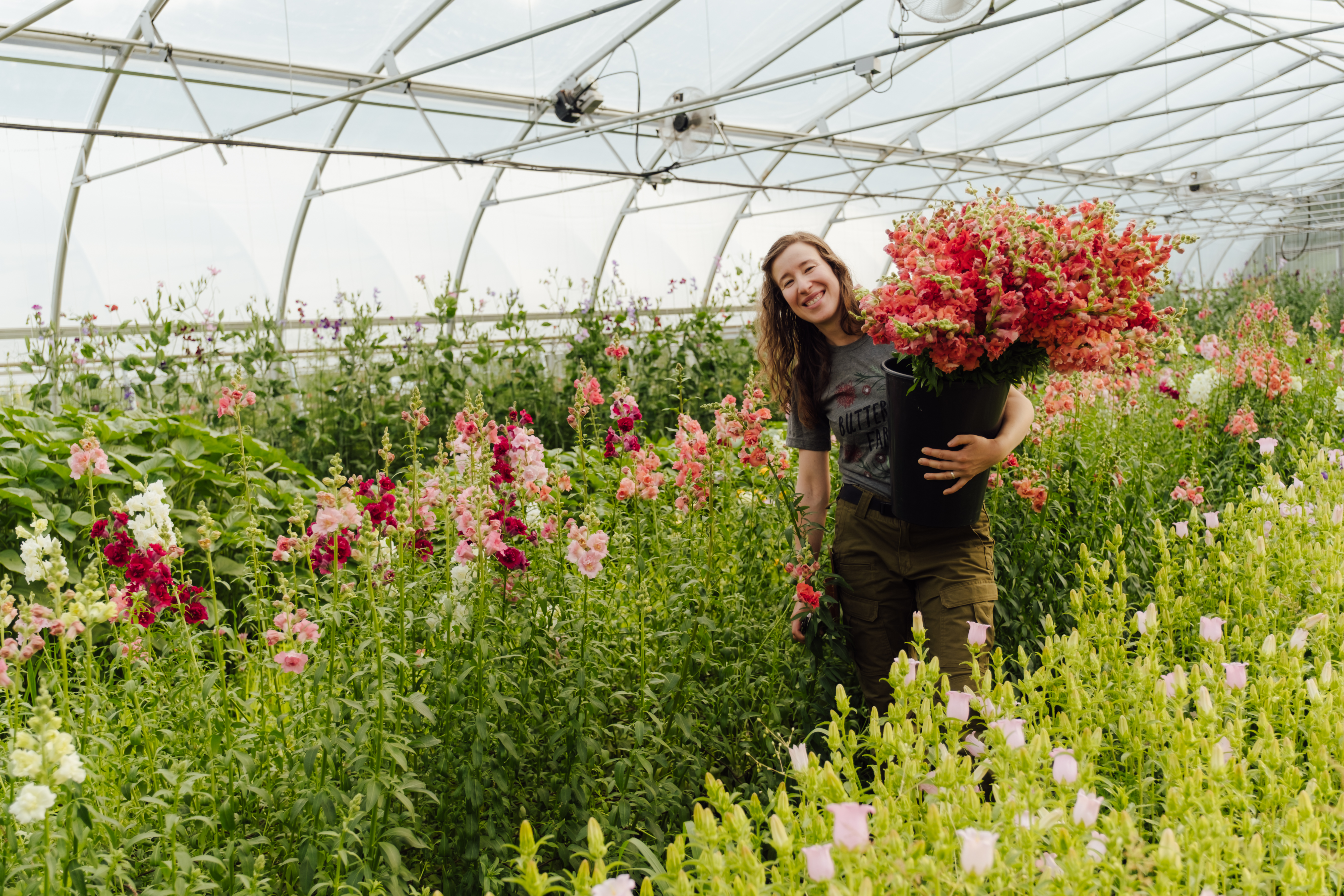 A woman with red hair holds an armful of flowers while walking through a greenhouse.