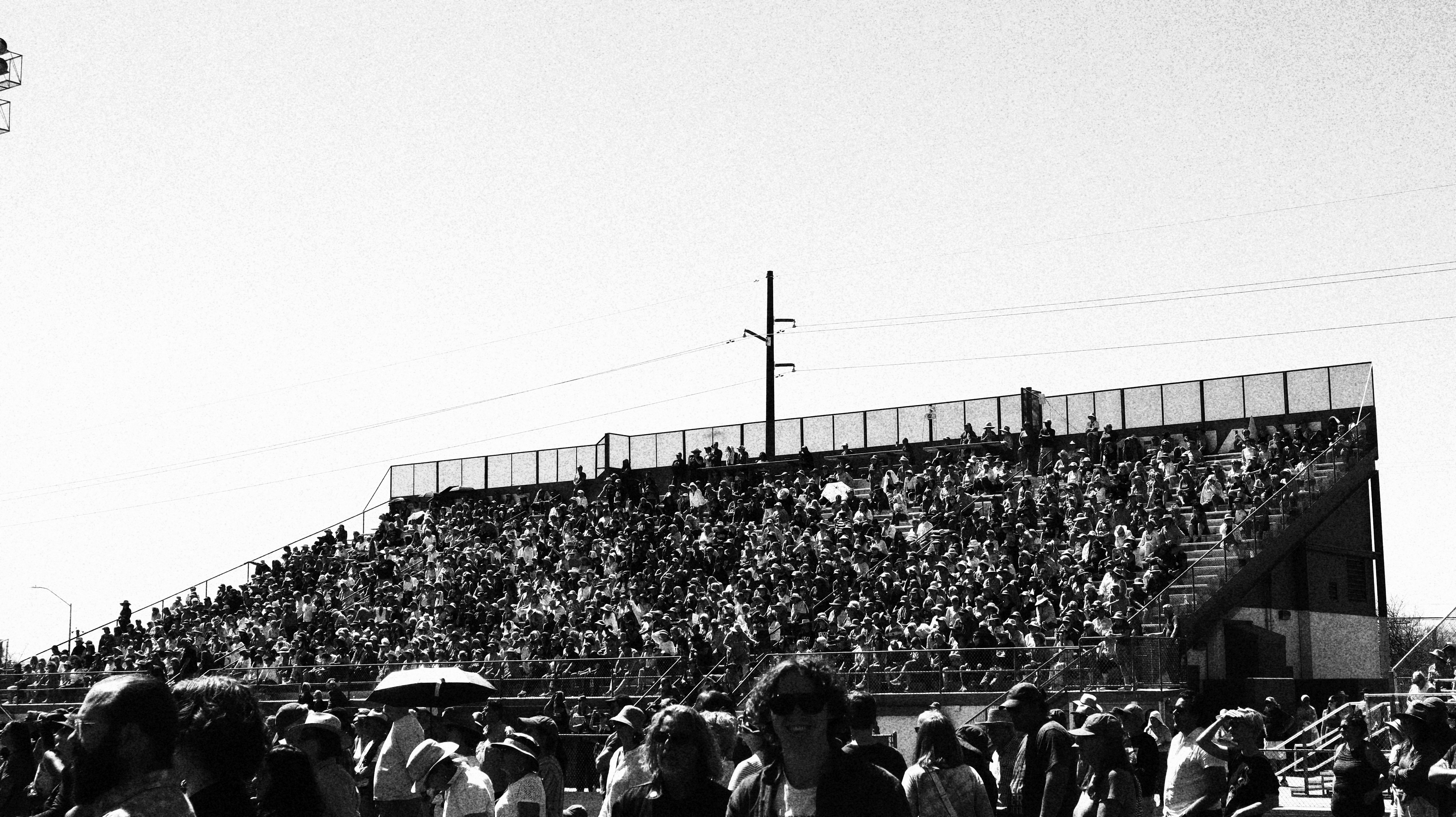 People sit in tiered bleachers and stand in the field in front of them.
