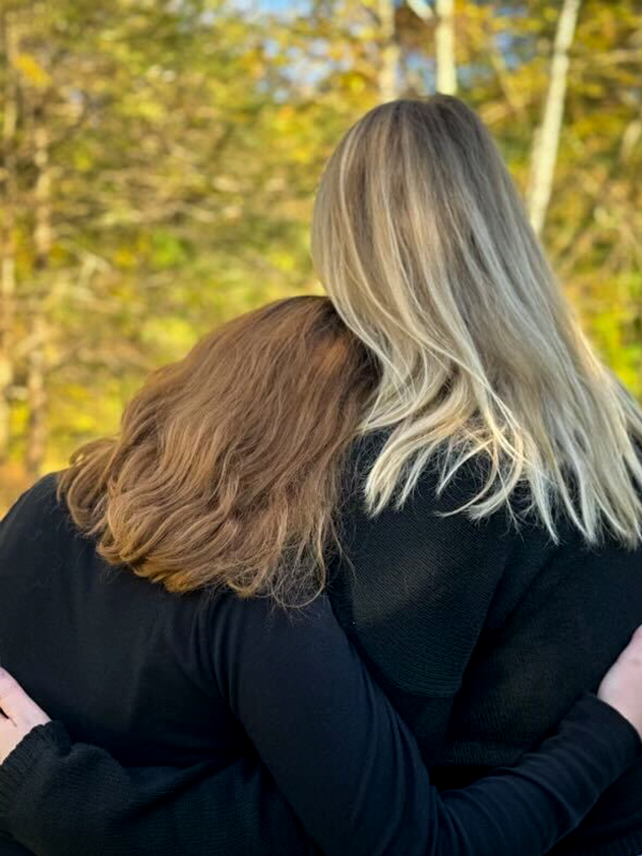 A long-haired mother and daughter embrace each other in a hug, facing away from the camera so their faces are not visible.