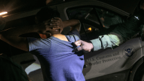 A photo of a young male person in a blue t-shirt with his hands behind his head. He stands in front of a white vehicle with the words "U.S. Customs and Border Protection" written in blue on one of the doors beside a circular seal with an eagle. A weathered male hand lit by a flashlight in the dark night grips the back of the young person's shirt.