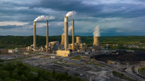 An aerial view of a power plant, which has smoke stacks, cooling stacks, and a heap of coal outside the facility. Though the sky is mostly cloudy, a beam of sun lights up one side of the power plant, which sits against a horizon of trees in Poca, West Virginia.