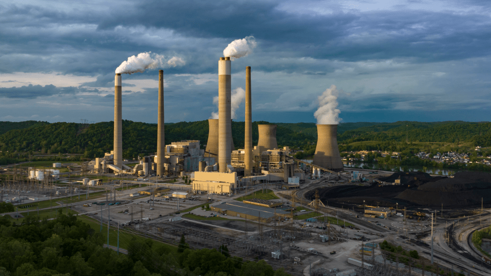 An aerial view of a power plant, which has smoke stacks, cooling stacks, and a heap of coal outside the facility. Though the sky is mostly cloudy, a beam of sun lights up one side of the power plant, which sits against a horizon of trees in Poca, West Virginia.