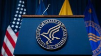 A podium which says "Health and Human Services" on it. There are four flags, including an American flag, in the background.