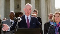 A balding man in a navy suit stands at a microphone on the steps of the US Supreme Court.
