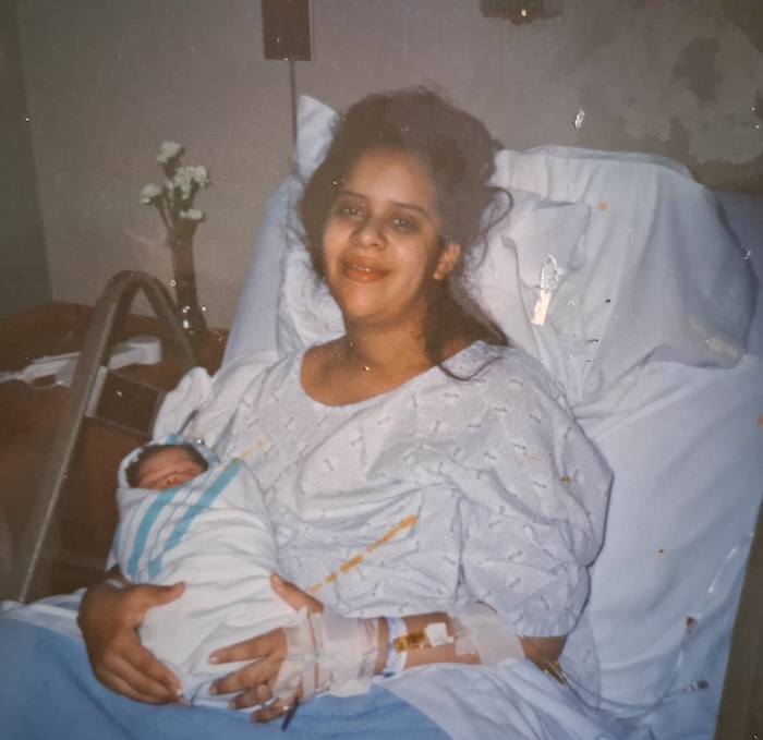 A light skinned black woman sits on a hospital bed with a baby wrapped in a blanket