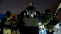 A group of ICE agents gathered in a circle, with the back of the uniform saying "Police Ice" being the most visible. It is evening in the photo
