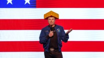 Elon Musk, wearing a cheese head, stands in front of an American flag.